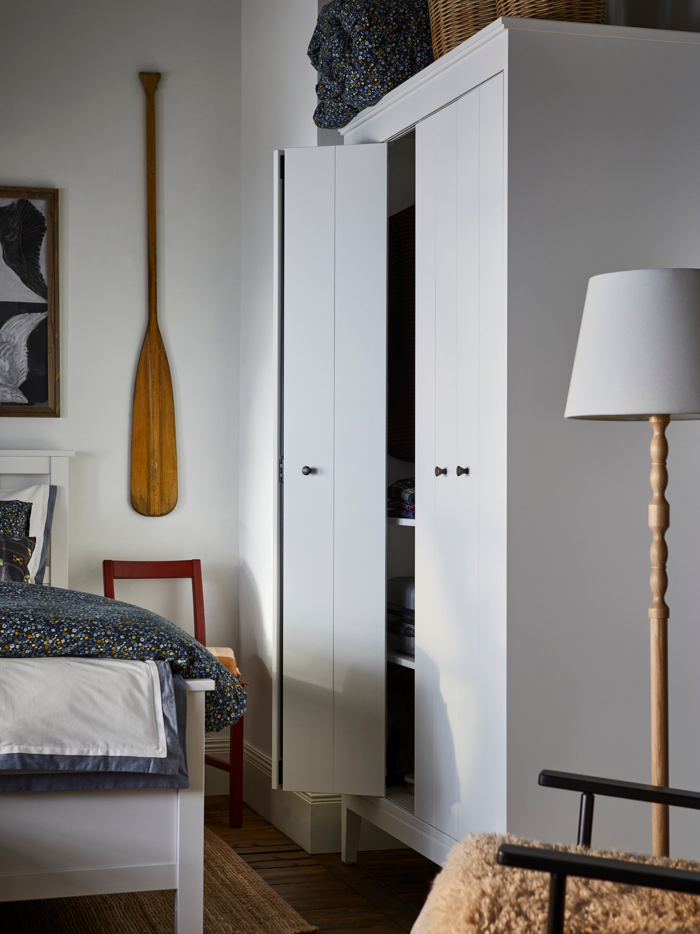 A white IDANÄS wardrobe stands in a corner of a bedroom near an ash/white KINNAHULT floor lamp and a HEMNES bed.