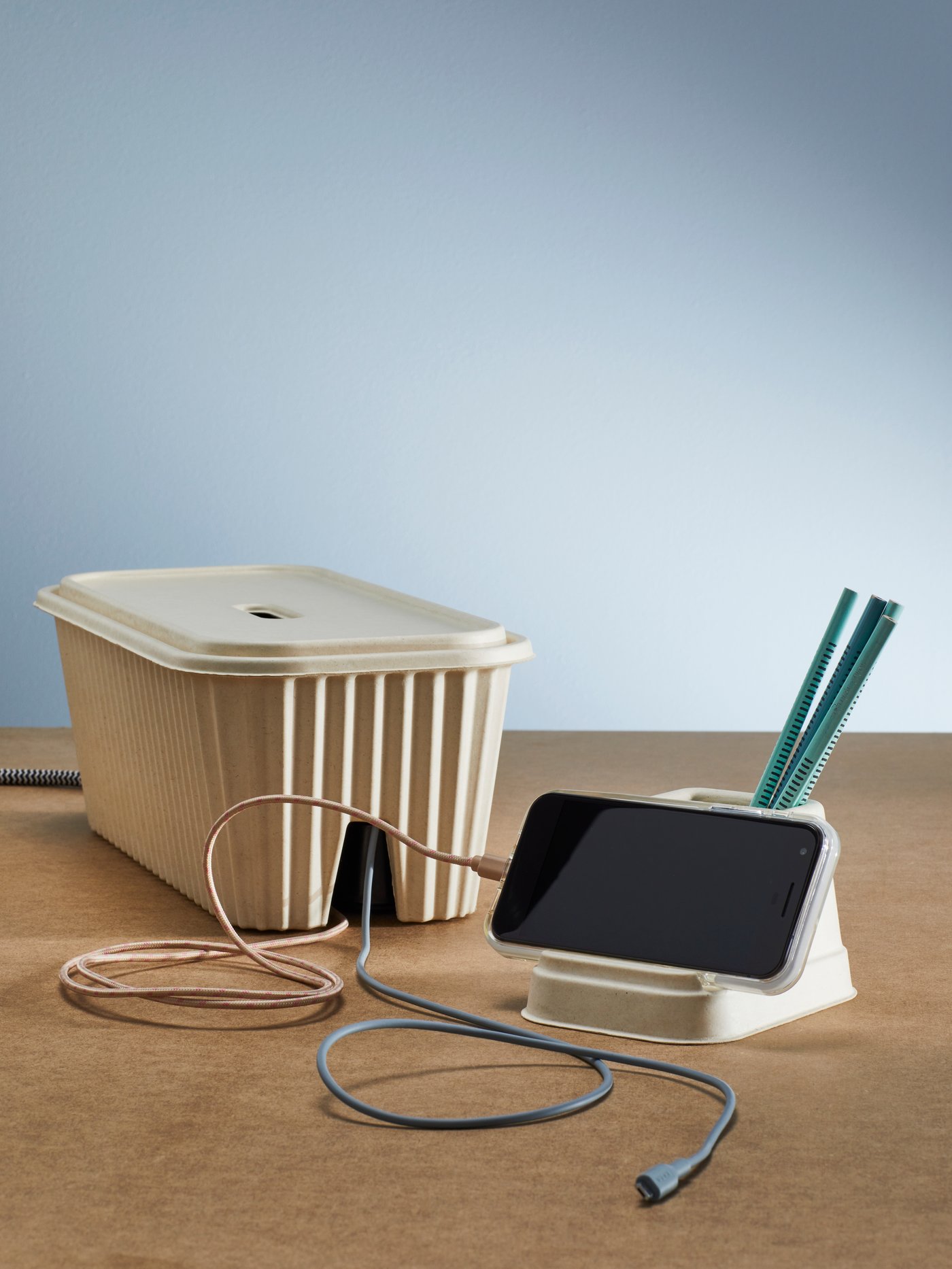A mobile phone is displayed on a natural-coloured HÖNSNÄT holder for mobile phone next to a HÖNSNÄT cable management box.