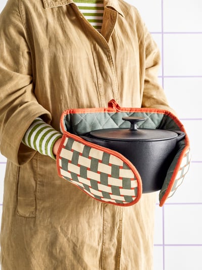 A person holding a black pot with lid while wearing a patterned RINNIG oven mitt in light beige/grey-green brown-red.