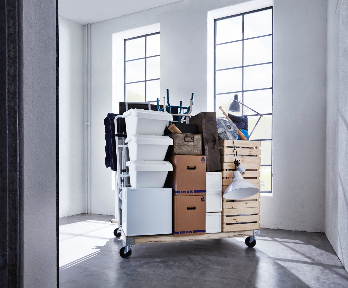 Eine Vielzahl von Umzugsutensilien und Kästen auf einer Holzpalette mit Rollen in einem leeren Zimmer.