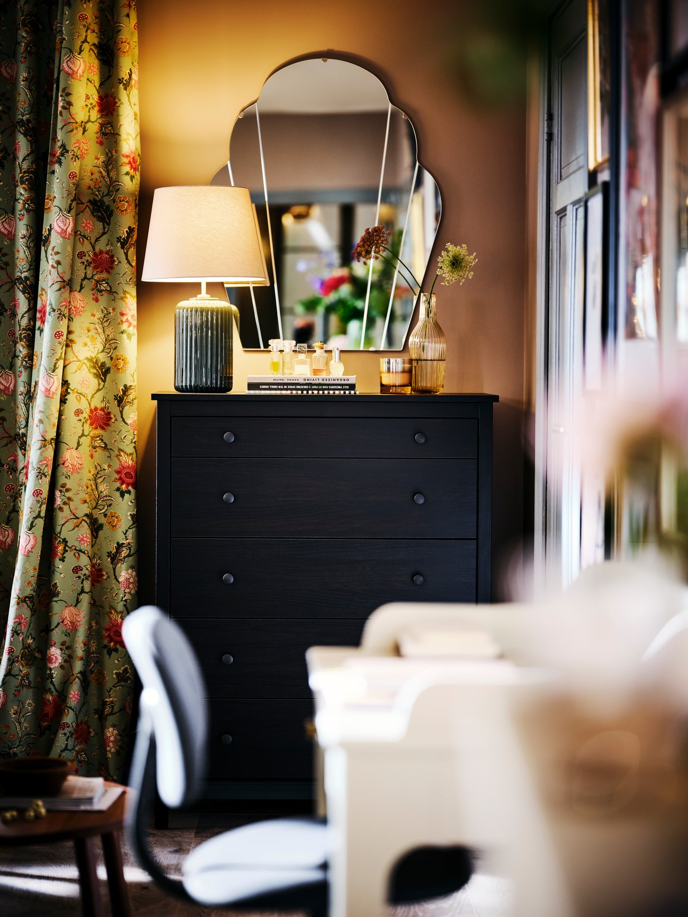 Una cómoda KOPPANG, con una lámpara de mesa SNÖBYAR encendida, contra una pared con un espejo LOMMARYD.