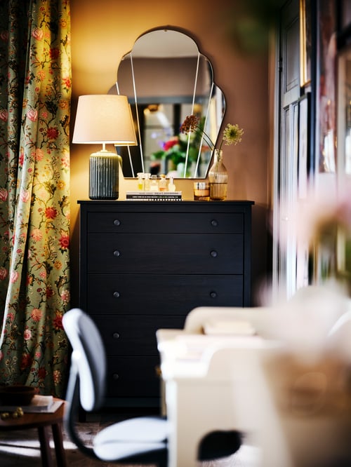 A KOPPANG chest of drawers, with a lit SNÖBYAR table lamp against a wall with a LOMMARYD mirror.