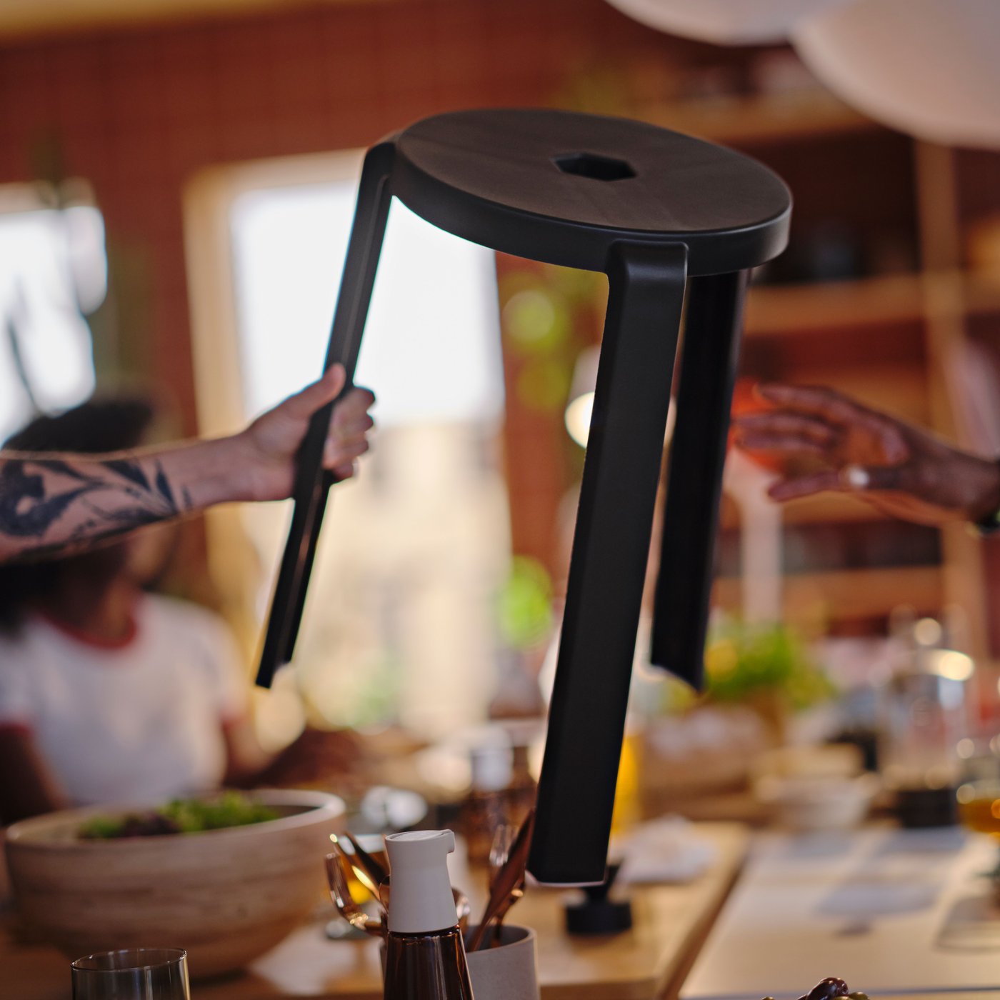 A tattooed arm is holding a dark grey IBBARBO stool in the air, reaching it over to a hand that is about to grab it.