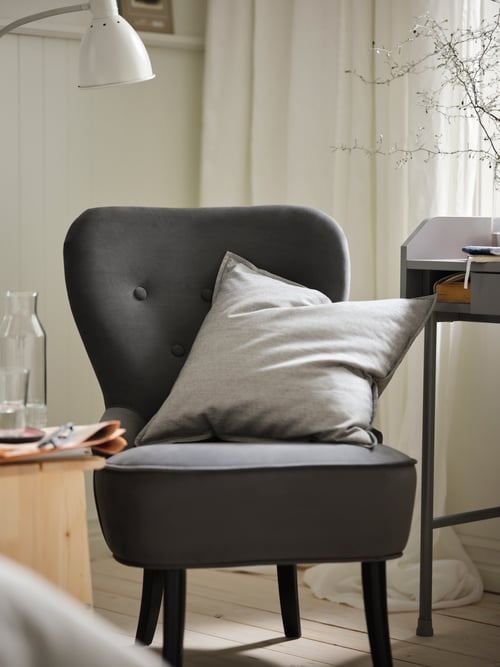 Living room shot of a grey armchair with a square cushion, and a rectangular desk to the side. A lamp is above the armchair.