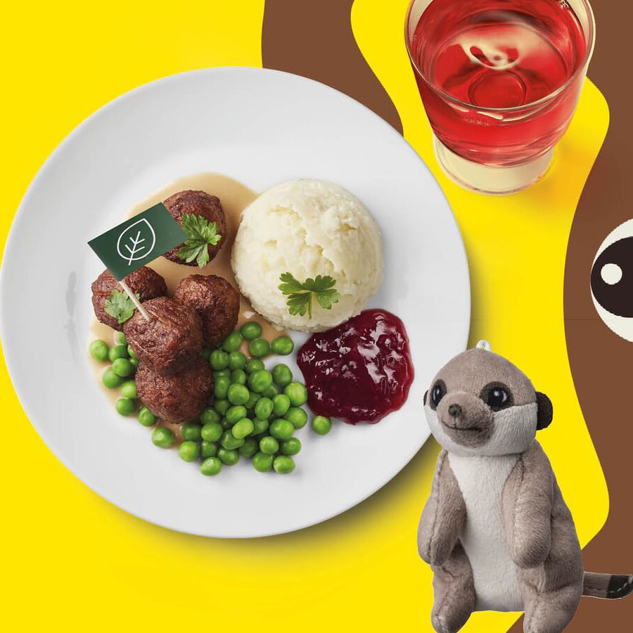 Auf einem gelben Hintergrund ist ein Kindermenü mit Fleischbällchen, Kartoffelpüree, Erbsen und Preiselbeeren sowie ein rotes Getränk und ein Kuscheltier dargestellt.