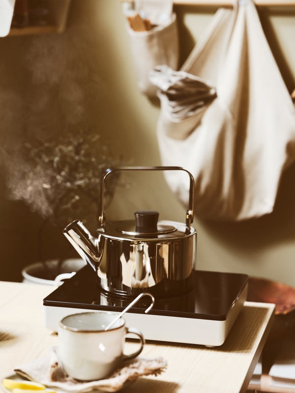 A kitchen worktop with a brewing teapot on top of a TILLREDA portable induction hob, next to a mug with a tea infuser.