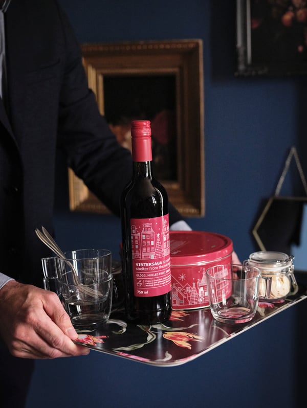 A man holds a flower-patterned tray with VINTERSAGA glögg, biscuits, glasses and almond flakes in a jar on it.