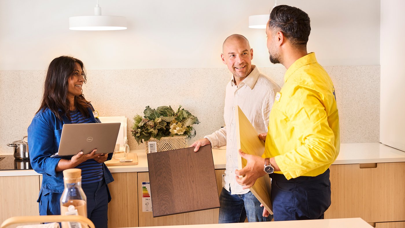 Konsultant IKEA w żółtej koszulce i pracowniczka IKEA w niebieskiej koszuli prowadzący usługę planowania kuchni w domu klienta. Obok konsultanta stoi klient trzymający front kuchenny w ręku.