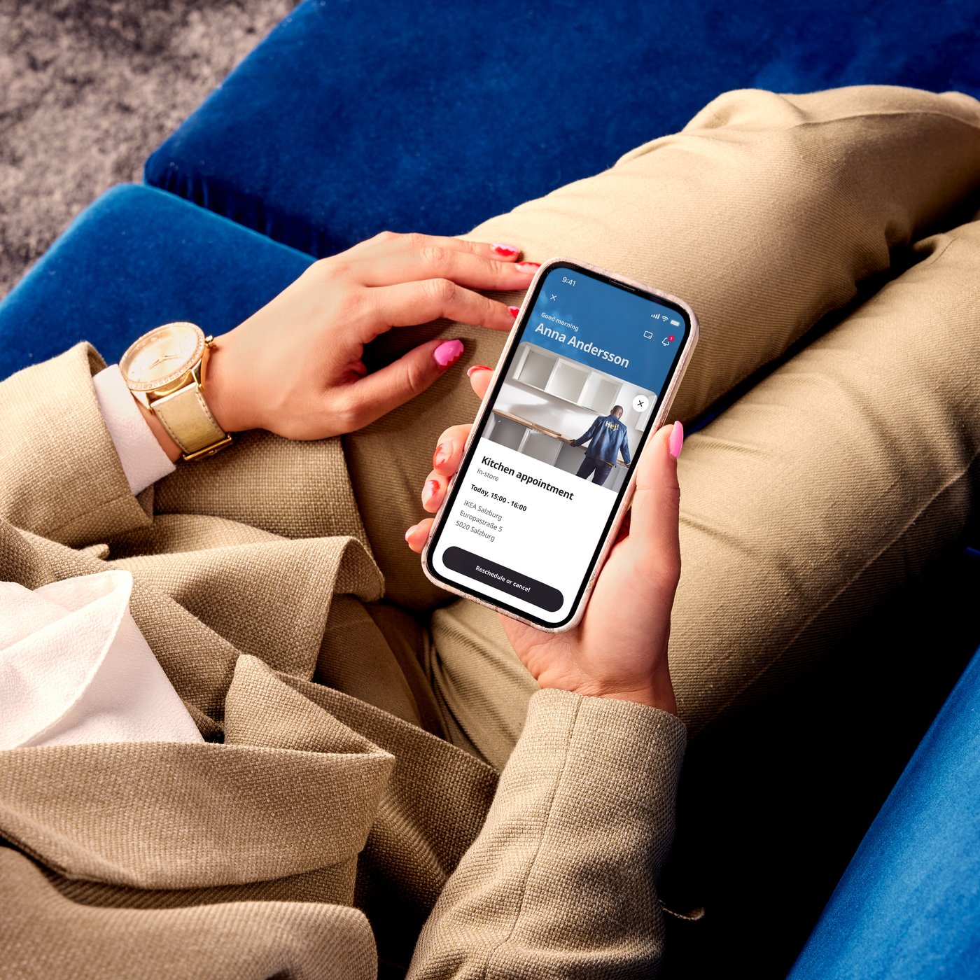 A person sitting on a blue sofa holds a smartphone displaying the IKEA app.