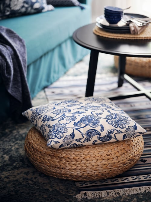 Un coussin avec une housse JUNIMAGNOLIA posé sur un tabouret en fibre de banane ALSEDA près d’une table basse KRAGSTA noire.
