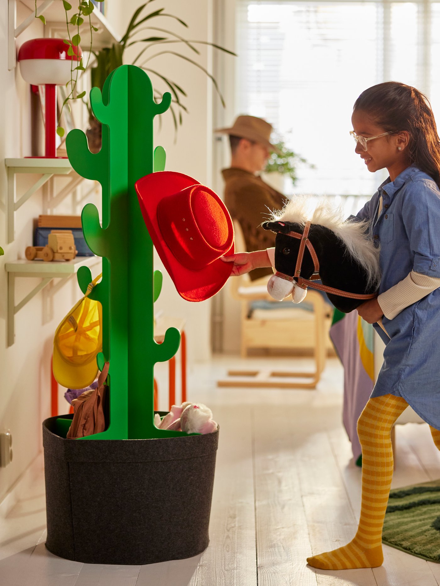 I ett solbelyst vardagsrum hänger ett barn på en käpphäst en röd cowboyhatt på ett kaktusformigt GREJSIMOJS klädställ.
