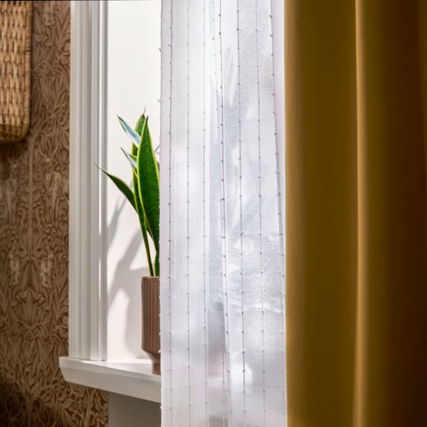 MATILDA sheer curtain in white hanging on window, with sunlight filtering through next to yellow curtain and plant.
