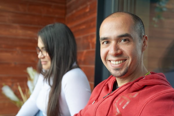 Edgar sitting on his balcony wearing a red hoodie smiling, his partner sits in the background.