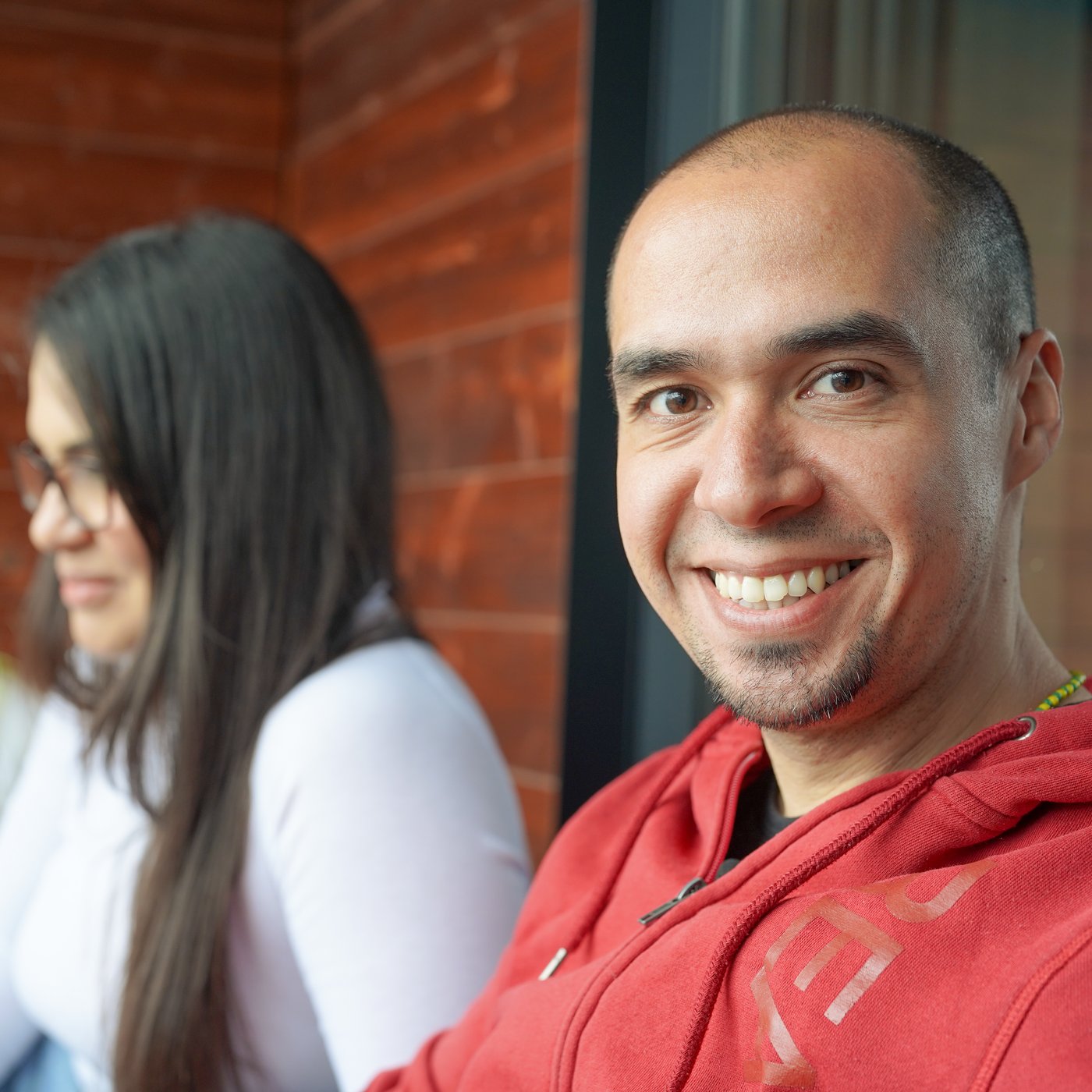 Eine Person sitzt in einem Raum und lächelt in die Kamera. Sie trägt ein rotes Hemd.