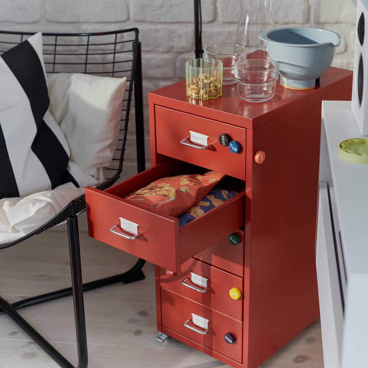 A room features a red HELMER drawer unit on castors, with four drawers filled with snacks, beside a modern chair.