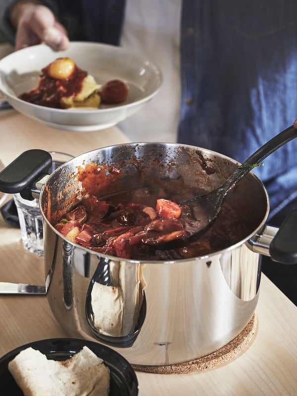 A person is using a black spoon to serve some stew out of a MIDDAGSMAT pot made of stainless steel into a white bowl.