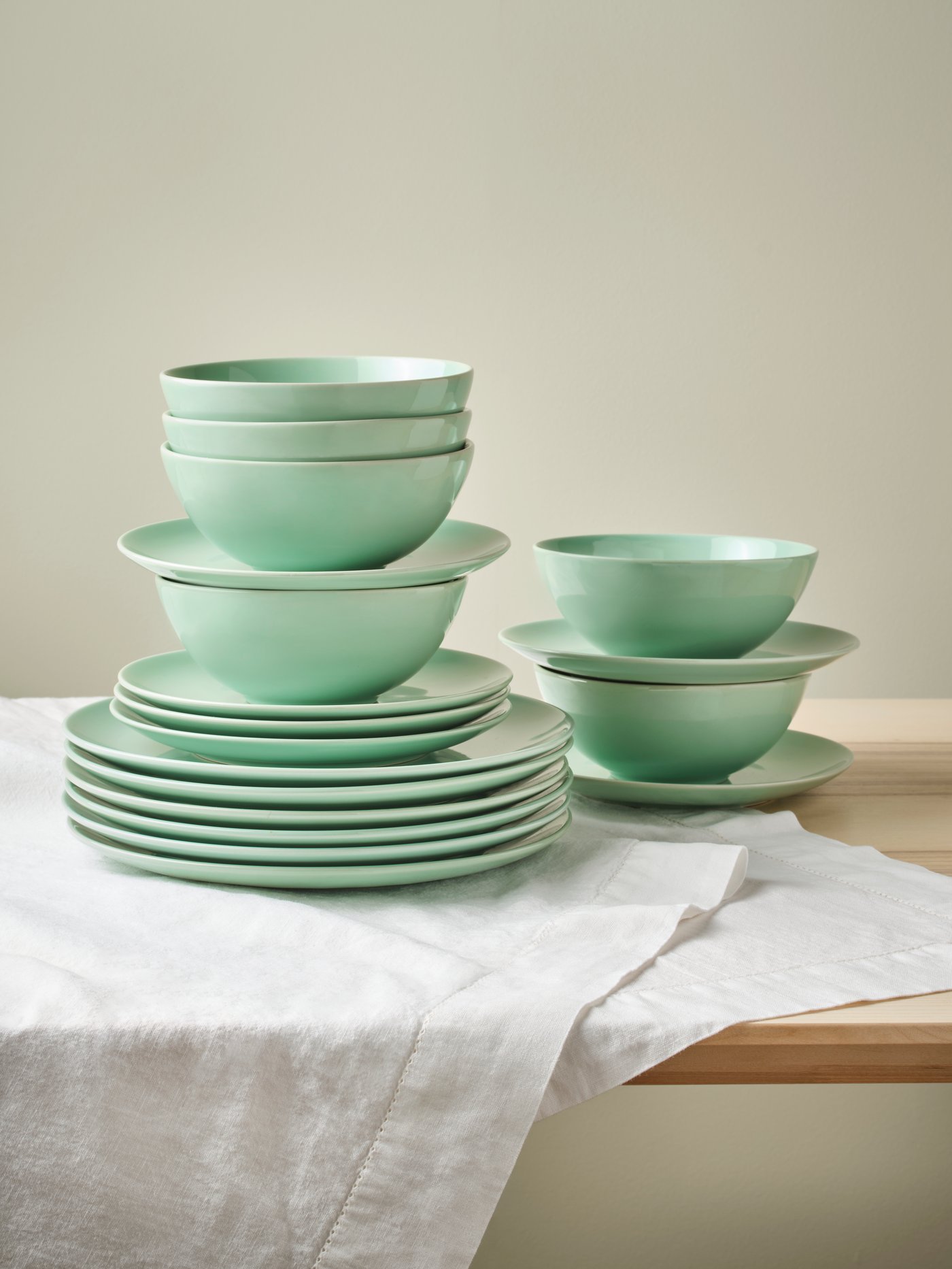Un service FÄRGRIK de 18 pièces, de couleur vert clair, comprenant des bols, des assiettes à dessert et des assiettes plates, est disposé en deux piles sur une table recouverte d'une nappe.