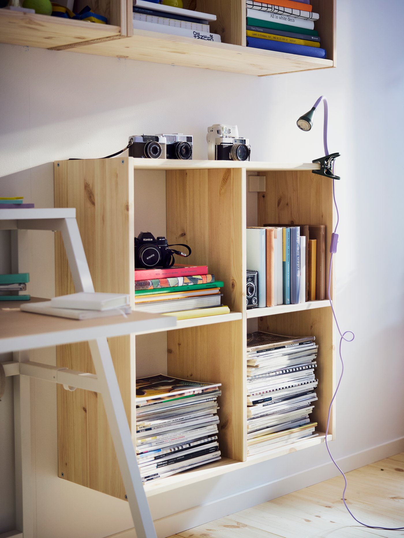 Närbild av IVAR hyllsystem i furu, upphängt på väggen bredvid ett skrivbord. Lådorna är fyllda med tidningar och kameror.