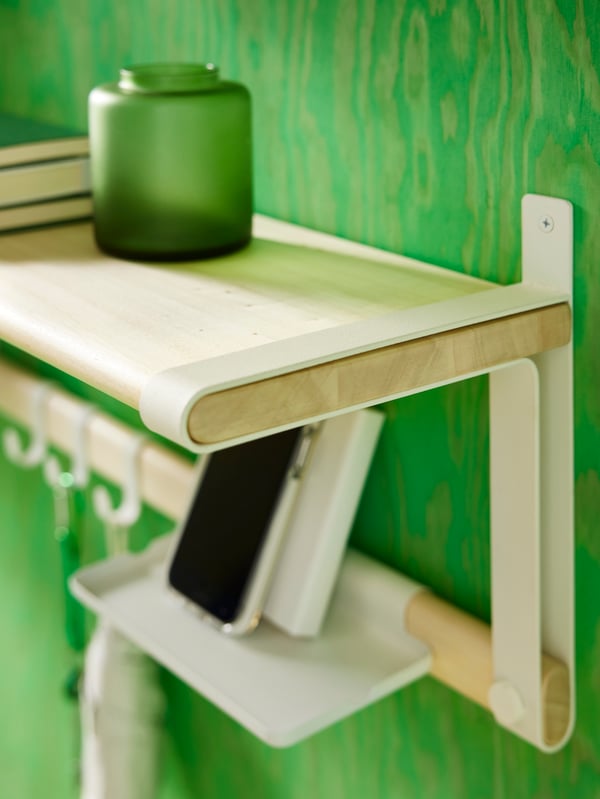 A SKOMAKARE wall shelf in white and aspen mounted on a green-colured wall, holding a vase, books and a mobile phone.