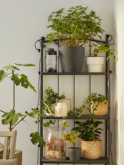 A grey, outdoor LÄCKÖ shelving unit is secured to a balcony wall and holds many potted plants, a lantern and a watering can.