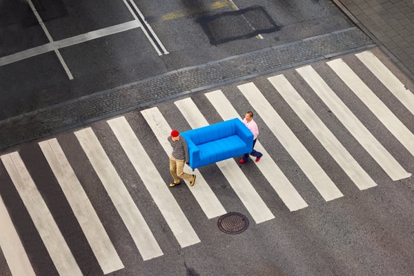 Two people carry a bright blue sofa across a black and white crosswalk on an asphalt street, seen from above.