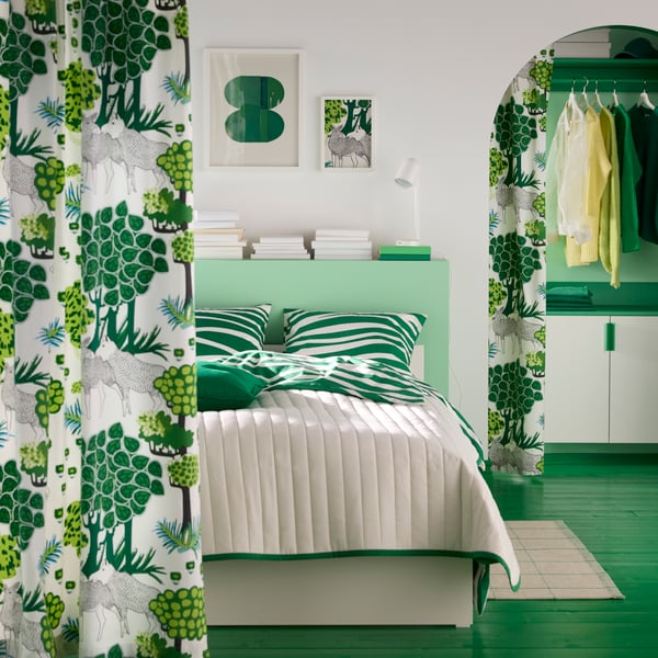 Een kamer met een bed met groene kussens en een wit dekbed, achter gordijnen van voorgeknipte ÄLGÖRT-stof met een bosmotief.