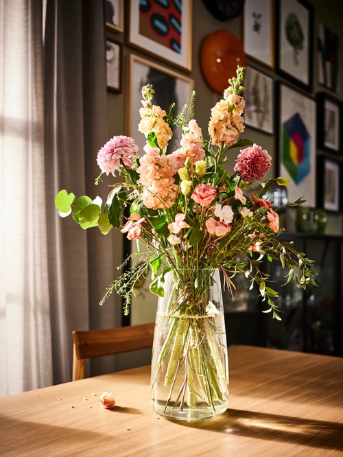 茎の長い花と植物の大きな花束がアレンジされた上品な背の高いガラス製花瓶があるリビングルームのテーブルのクローズアップ。