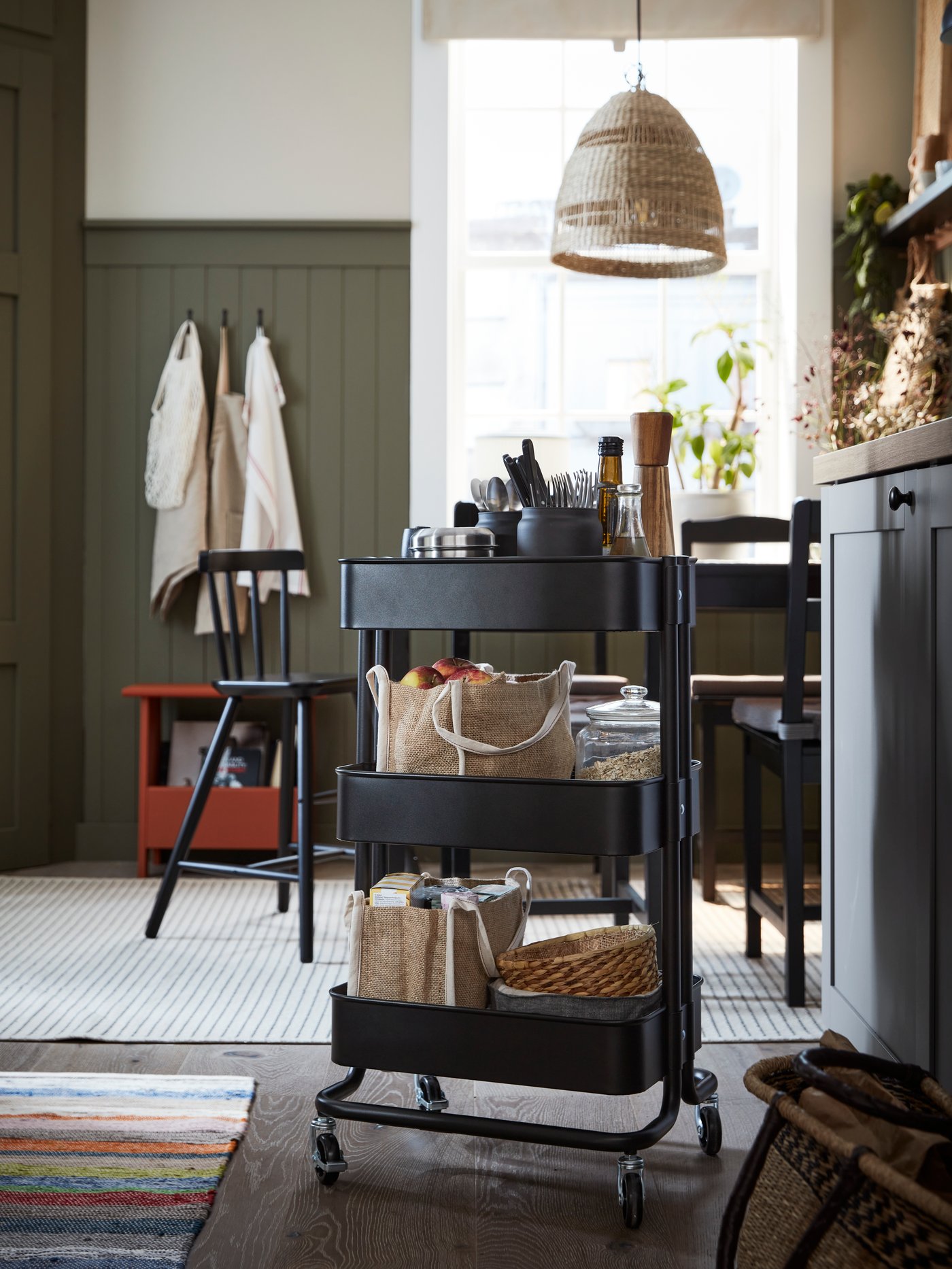 A black RÅSKOG trolley with many things on it including GULLRISMOTT food storage bags and food stands near a black table.