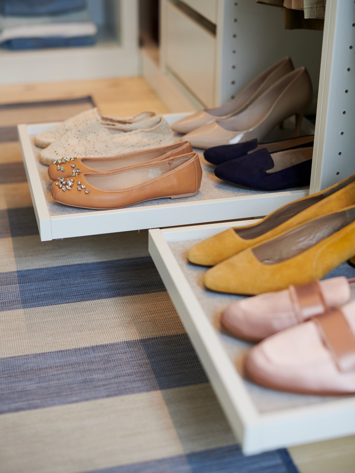 Närbild på utdragbara KOMPLEMENT skohyllor i en PAX garderob med flera par skor i olika färger och stilar, placerade på två hyllplan ovanför ett rutigt golv.