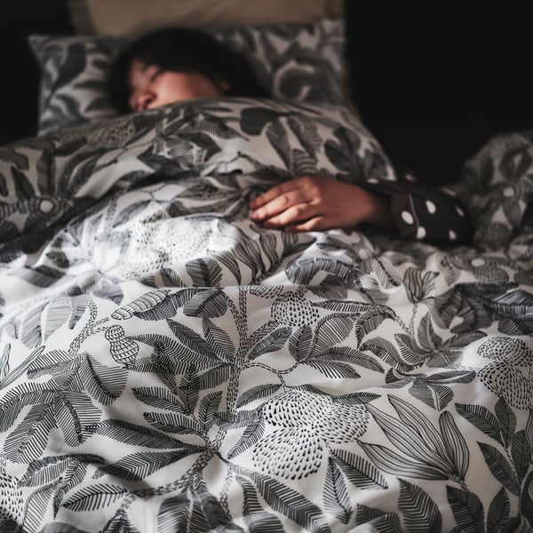 A person asleep in a SLATTUM upholstered bed under a duvet that’s covered in black and white BROKDRACENA bed linen.