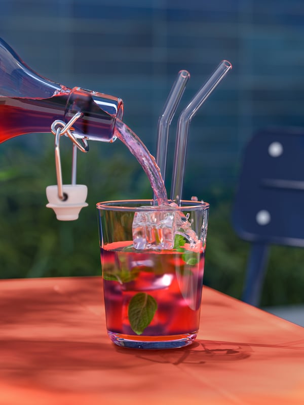 A clear glass IKEA 365+ glass with see-through drinking straws on a table is filled with a drink from a bottle with stopper.
