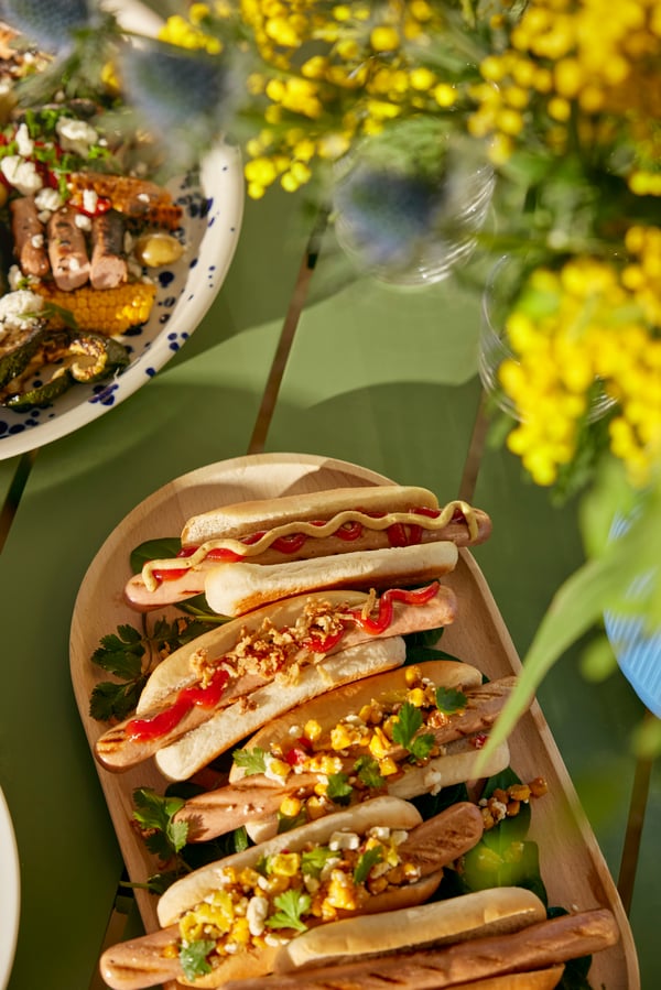 Various hot dogs with colourful toppings are laid out on a wooden tray, next to a flower arrangement on a green table.