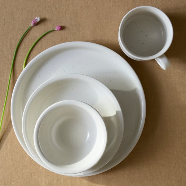 A set of DAGGASTER dinnerware in dotted/light beige, including plates and bowls, with a cup and some flowers on a table.