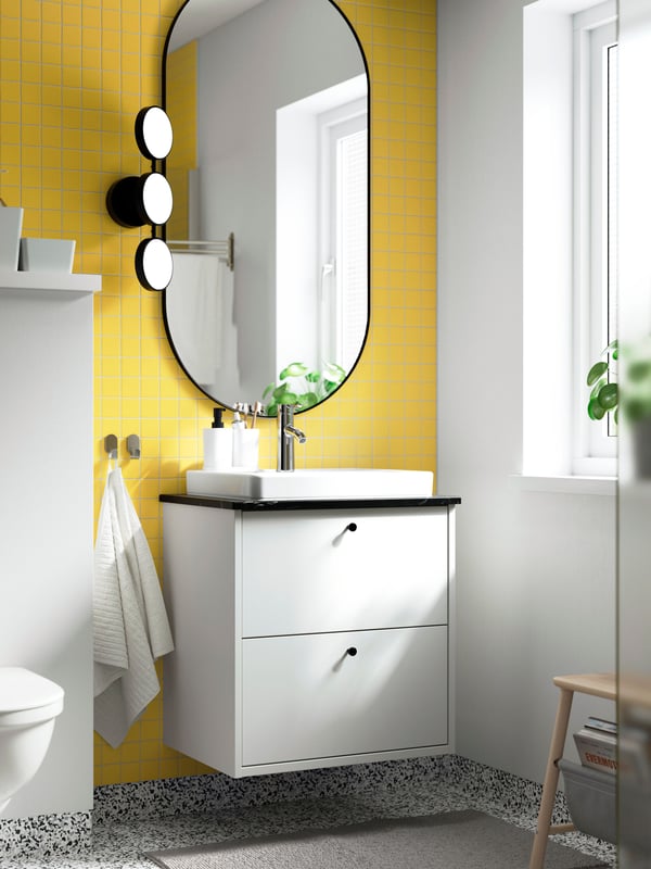 A white HAVBÄCK washstand is placed on a yellow wall below a black oval mirror and next to a black KAMBOMBA LED wall lamp.
