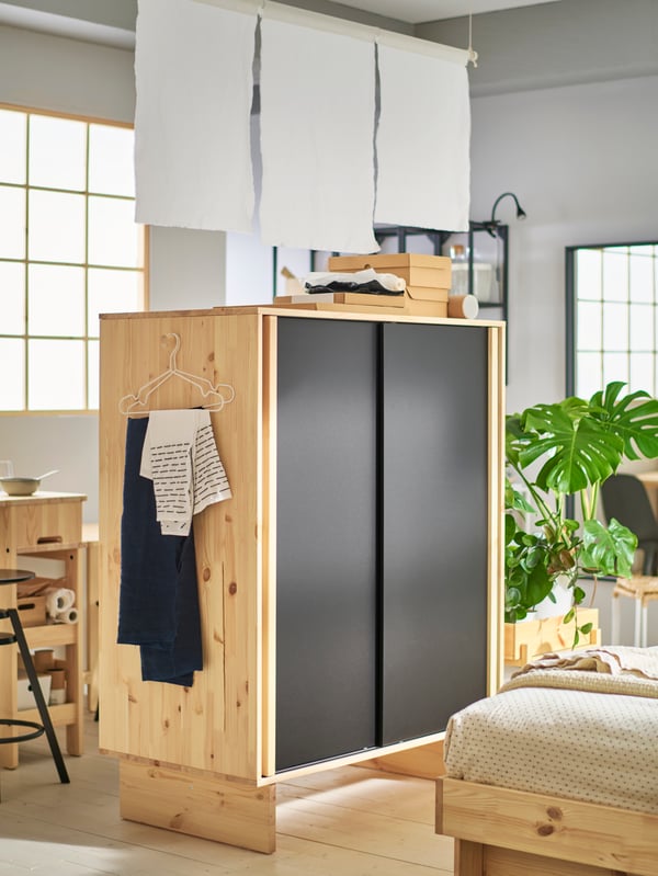 GLAMBERGET wardrobe in untreated wood with black sliding doors, placed in the middle of the room as a room divider.