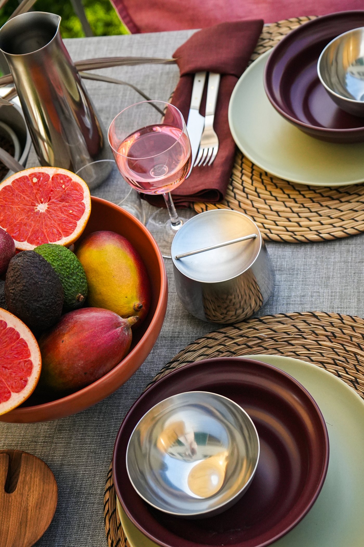 Décoration de table avec une corbeille à fruits BLOMVECKLARE, des pamplemousses, du vin rosé, des couverts enveloppés dans une serviette violette, des assiettes et de la vaisselle argentée pour un dîner raffiné.