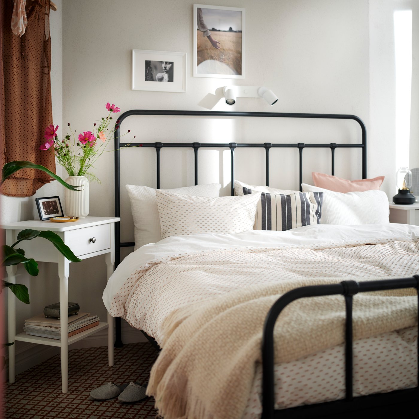 A black STJÄRNÖ bed covered with SCHERSMIN and BANDKNIPPE bed linen with a cushion in a KORALLBUSKE cover among the pillows.