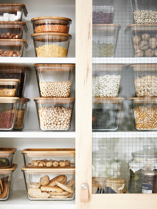 Küchenschrank mit Glastüren, in dem IKEA 365+ Vorratsbehälter mit Deckeln zu sehen sind, die mit Nüssen, Samen, Bohnen und anderen trockenen Vorräten gefüllt sind.