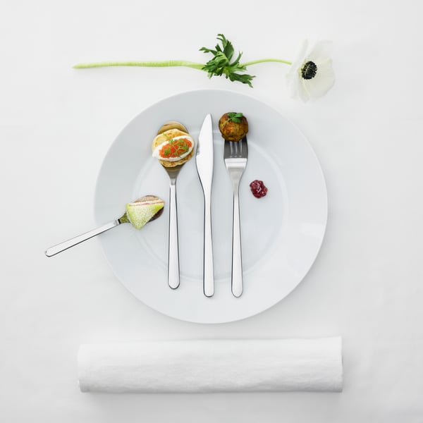 Un juego de utensilios FÖRNUFT encima de un plato blanco. Cada utensilio tiene una comida diferente, como pastel y caviar.