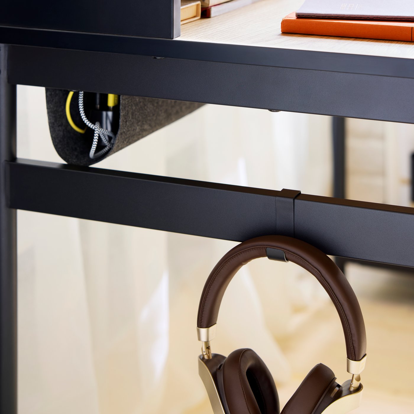A close-up of the hook on the side of a MULLSJÖ desk with headphones hanging on it and neatly wrapped USB cables above.