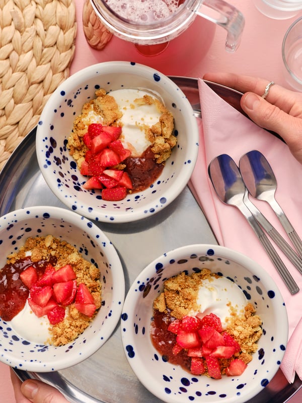 Une personne tient un plateau sur lequel sont posés trois bols de déjeuner garnis de biscuits KAFFEREP émiettés, de fraises et de confiture.