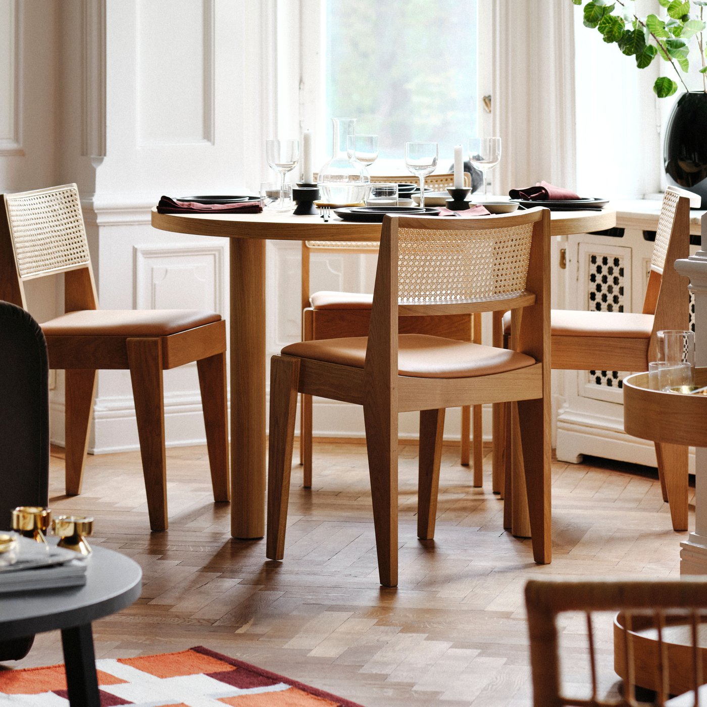 A living room dining set with an oak veneer STOCKHOLM 2025 table and oak and rattan chairs placed by large bay windows.