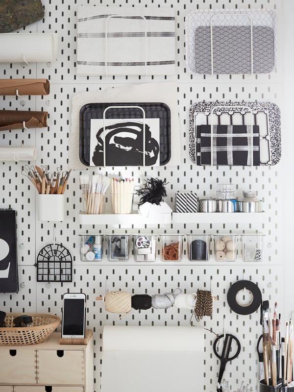 A white pegboard with organizers hanging on it with paintbrushes, pens, paper, art supplies, trays and more.