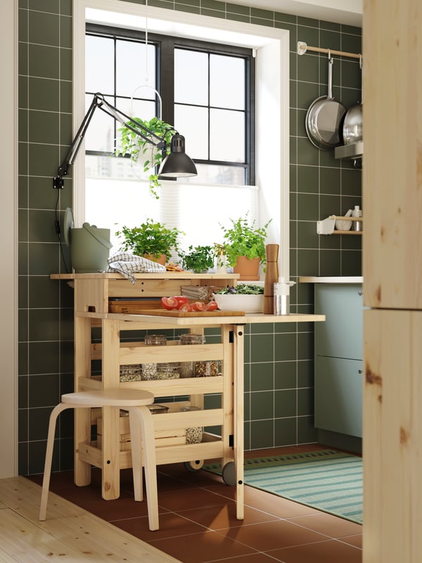 A RESARÖ mobile drop-leaf table with storage is standing in a kitchen, with kitchenware on top of it and a stool next to it.