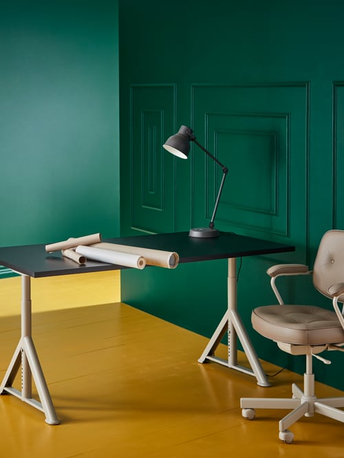 Workspace with green walls, desk with adjustable height, work lamp, adjustable office chair, pendant lamp shade, paper rolls.