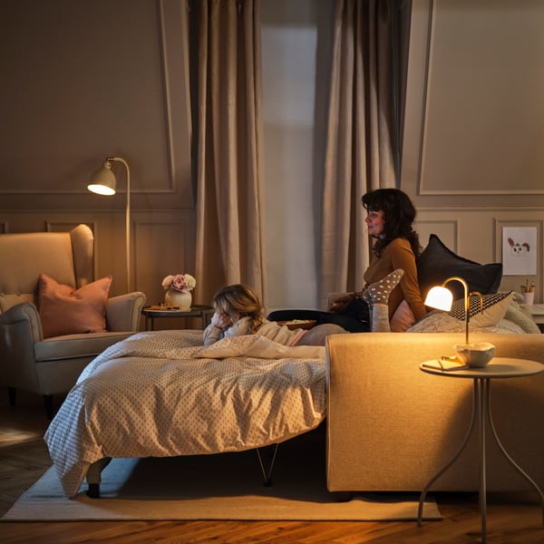 Um adulto e uma criança estão num sofá-cama EKHOLMA de 2 lugares, configurado como cama, junto a uma poltrona STRANDMON de abas e a um candeeiro.