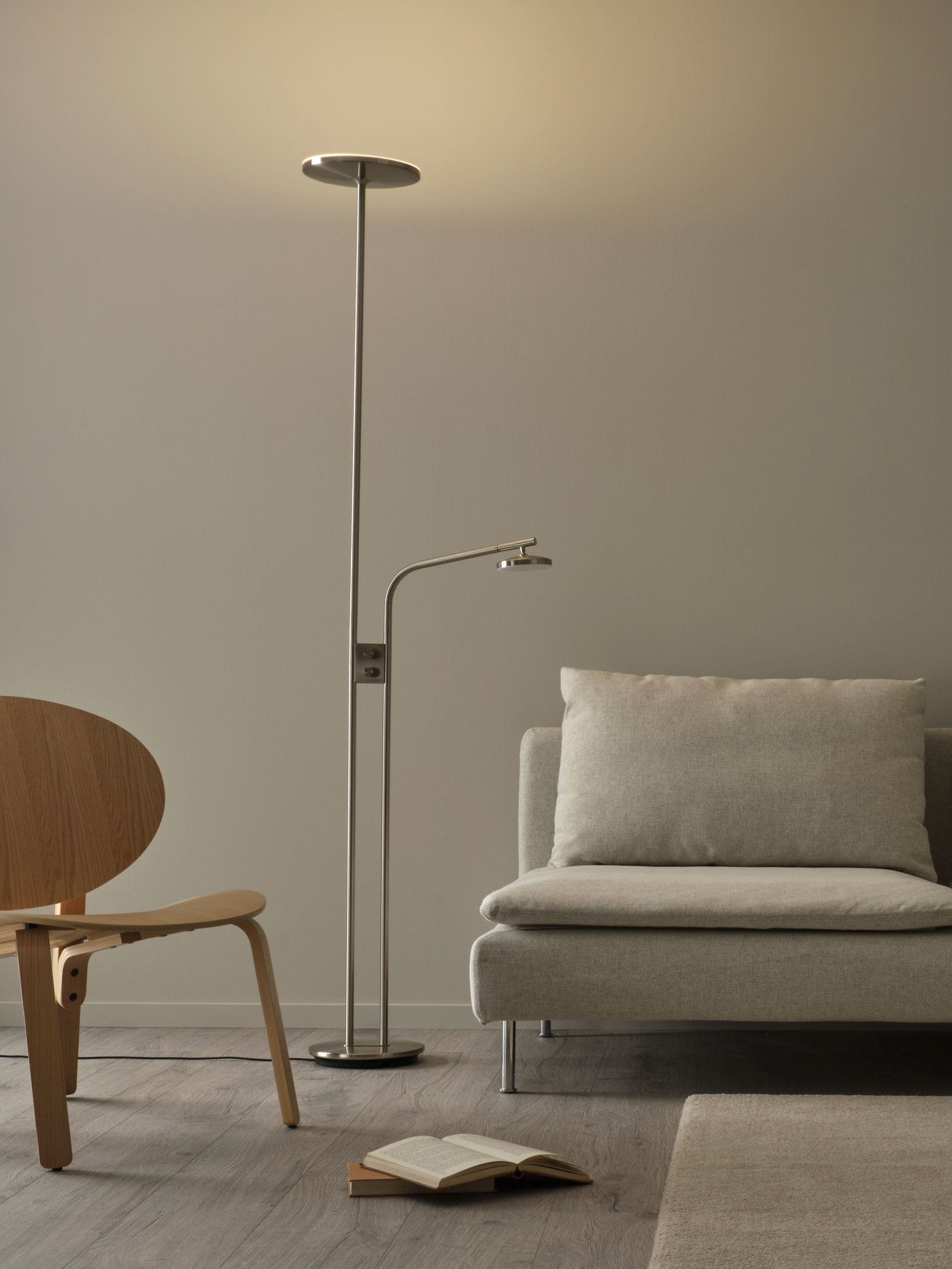 En dimbar ISJAKT LED-golvlampa i nickelplätering står mellan en beige soffa och en fåtölj i trä.
