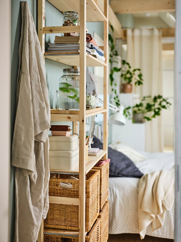 Es ist ein IVAR Regal neben einem Bett mit vier BRANÄS Körben aus Rattan zu sehen, die auf zwei der unteren Regalböden stehen. Im übrigen Regal befindet sich eine Vielzahl an Gegenständen.