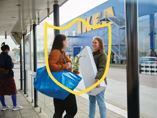 Two women standing and talking outside the entrance to an IKEA store. One is holding a blue FRAKTA shopping bag with a potted plant, the other is holding a white flat-pack.