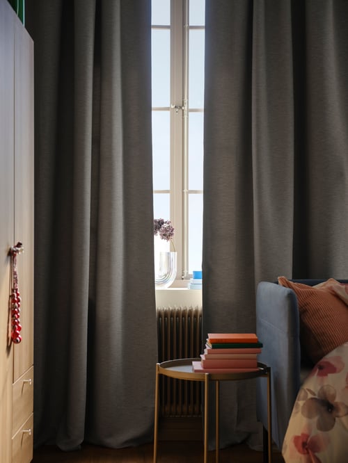 A pair of grey VILBORG room darkening curtains partially cover a window. In front of the window is a round GLADOM tray table.